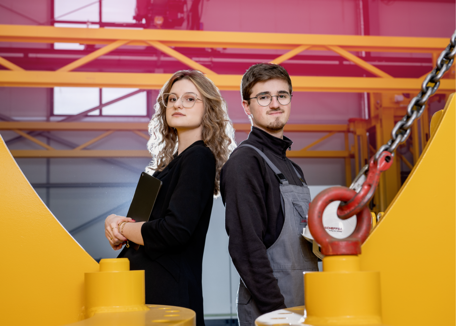 Eine Frau und ein Mann stehen Rücken an Rücken in der Produktionshalle von Scheffer Krantechnik. Im Hintergrund erkennt man einen gelben Kranteil von Scheffer.